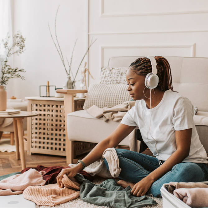 A woman with braided hair sits on a white sofa in a bright living room, wearing white headphones and sorting through a pile of folded clothes on the floor. She is dressed in a white ribbed t-shirt and blue jeans.