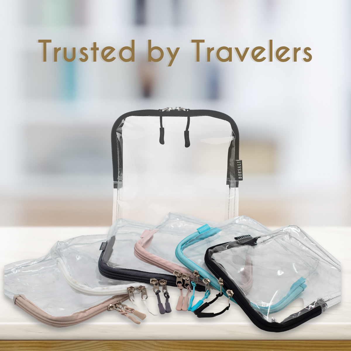 A set of clear plastic zippered travel pouches with black, pink, and blue handles, arranged on a light wooden surface with a blurred background and the text 'Trusted by Travelers' at the top.
