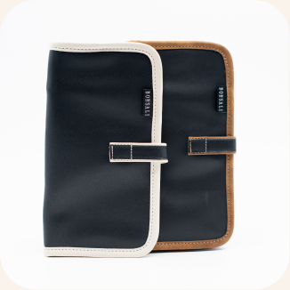Two slim folio-style travel wallets or organizer cases shown overlapping on a light gray background. The front case is dark navy with cream trim and a navy strap closure; the rear case is dark navy with tan trim and a tan strap closure.