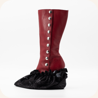 A tall red knee-high boot with silver metal studs down the side and a black ruffled pointed-toe slipper underneath, shown on a pale pink background.